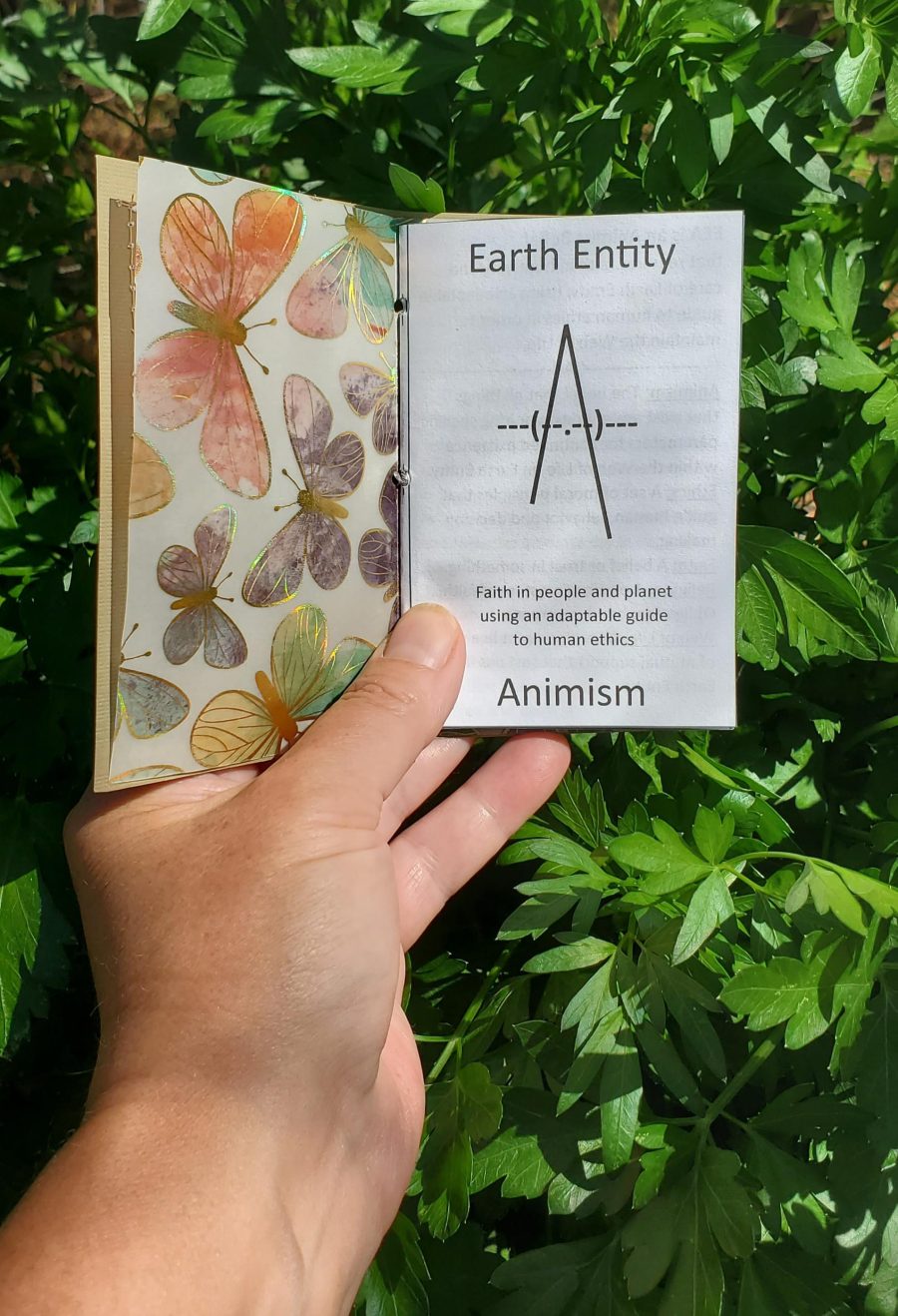 A picture of a human hand holding a small booklet open in front of parsley in the garden.  The first page of the booklet is displayed showing the title page of the signature (stack of pages) containing the tenant of Earth Entity Animism.